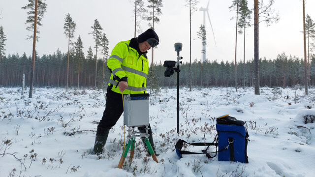 Tutkimustyöntekijä käyttää kolmijalkaisen jalustan päälle asetettua painovoiman mittauslaitetta talvisessa maastossa.