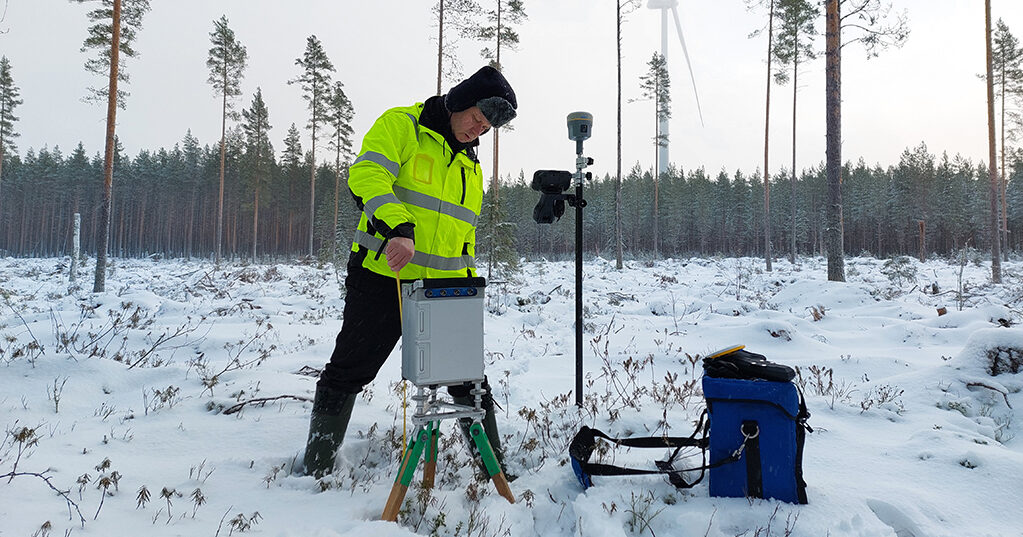 Tutkimustyöntekijä käyttää kolmijalkaisen jalustan päälle asetettua painovoiman mittauslaitetta talvisessa maastossa.