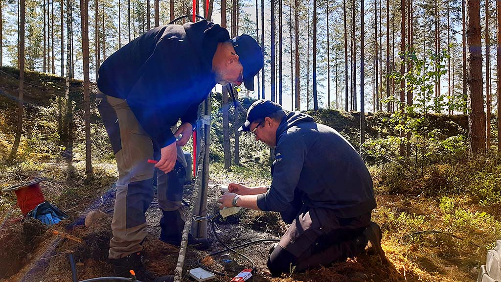 Kaksi tutkijaa suorittamassa geoenergian mittausta maastossa. Aurinkoinen syyspäivä metsän keskellä.