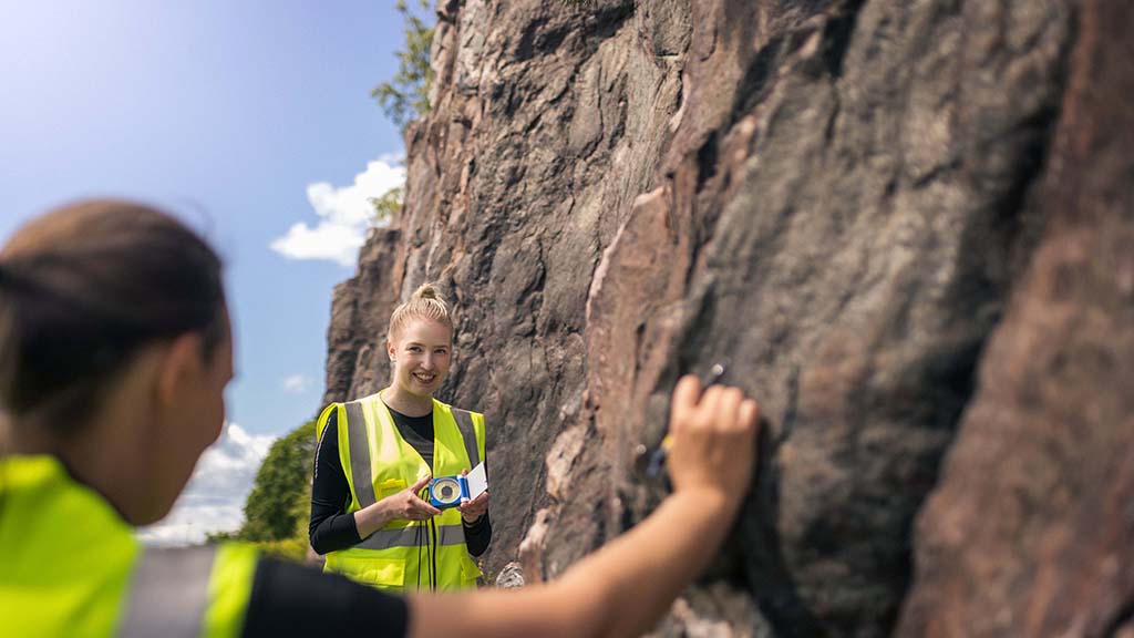 Hymyilevä GTK:n tutkija kallioseinämän vieressä mittaamassa kallioleikkauksen rakennesuuntia yhdessä kollegansa kanssa. Aurinkoinen kesäpäivä.