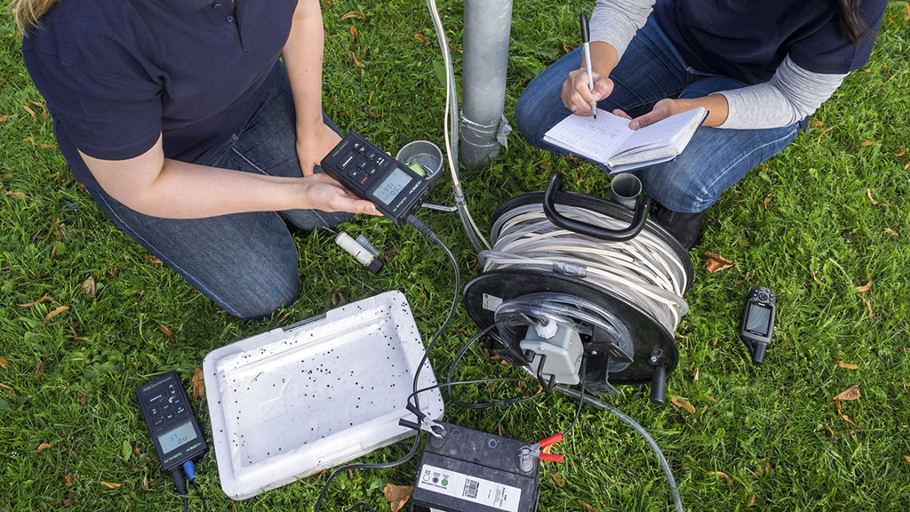 Kaksi GTK:n tutkijaa ottamassa maaperän vesinäytettä ruohikkoisella kentällä. Toinen tutkijoista merkitsee mittaustuloksia vihkoon.