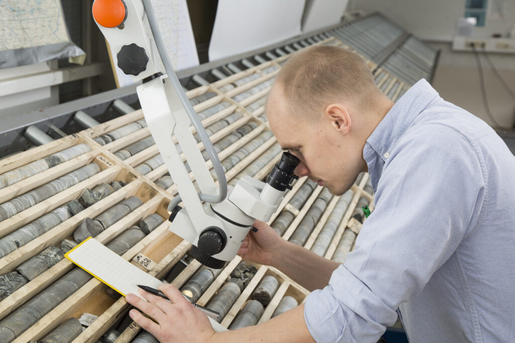 A person studies bedrock samples, drill cores, with a microscope. The samples are in a wooden table.