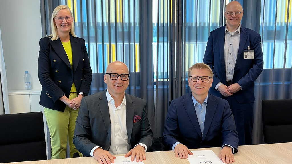 There are four persons smiling to the camera. In front 2 persons have just signed a contract and are sitting on the table. Behind them are two persons standing.