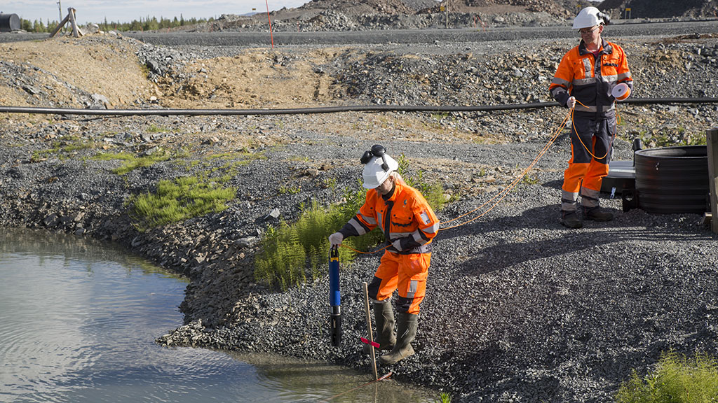 Kaksi tutkijaa ottamassa näytettä kaivosalueen vesialtaasta, taustalla kaivoksen sorakumpareita. 