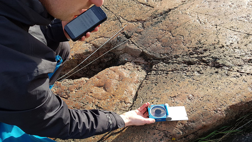 Tutkijan kädessä mittauslaite, jolla hän tutkii kallioperää merenrannalla aurinkoisena päivänä.