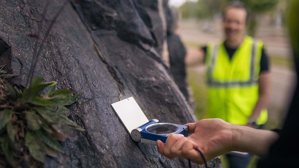 Kaksi tutkijaa kallioseinämän vieressä mittaamassa kompassilla kesäisenä poutapäivänä.