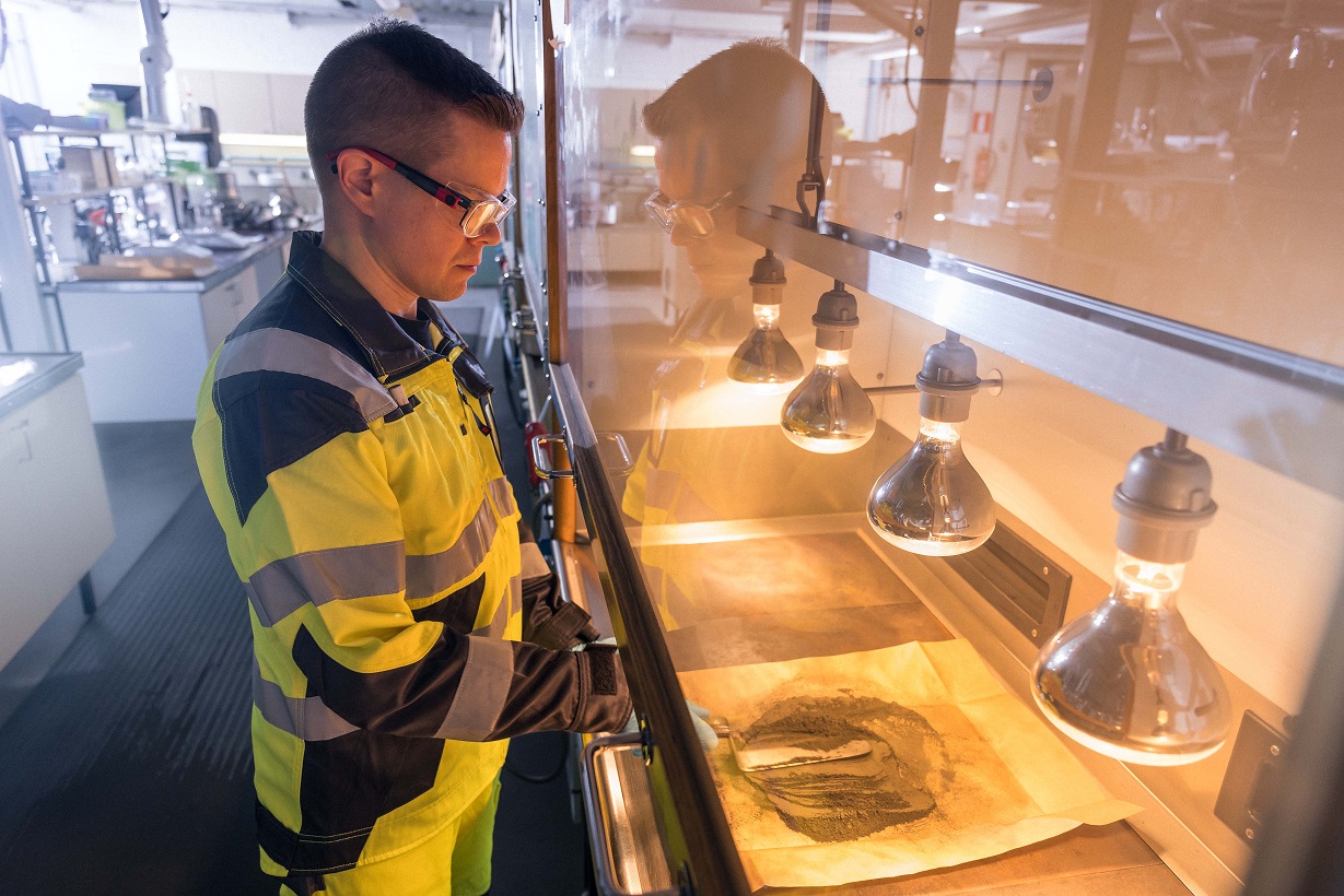 A person is drying a sample with a spatula. The sample is behind a screen and there are heat lamps to dry the sample.