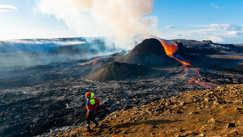 Kuvassa on purkautuva tulivuori. Etualla kävelee tutkija suojavarusteissa.