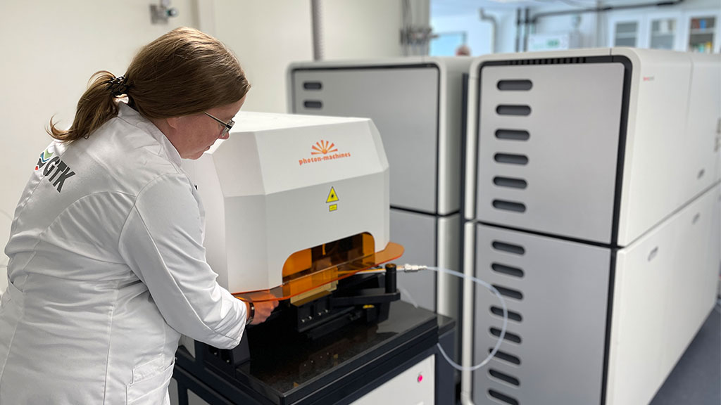 A person in a laboratory coat in front of a large laboratory equipment. She is placing a sample to it.