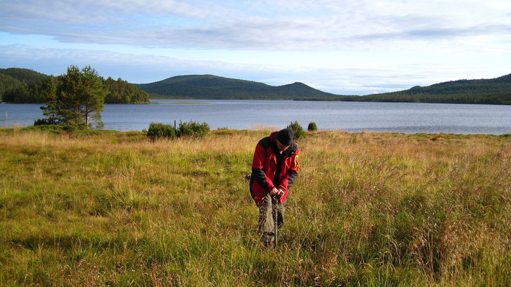 GTK researcher taking a soil sample in Northern Finland, with a lake and fells in the background.