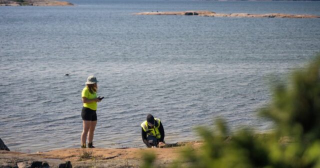 Kaksi henkilöä keltaisissa huomiovaatteissa kalliolla ottamassa näytteitä. Taustalla meri.