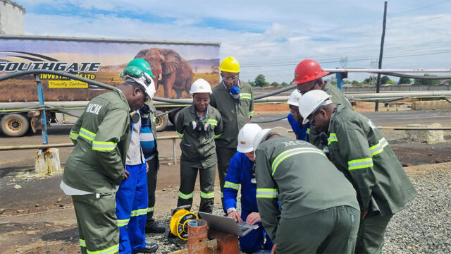 A group of people in protective clothing looks at a laptop outdoors at a worksite. A truck and pipes are in the background.