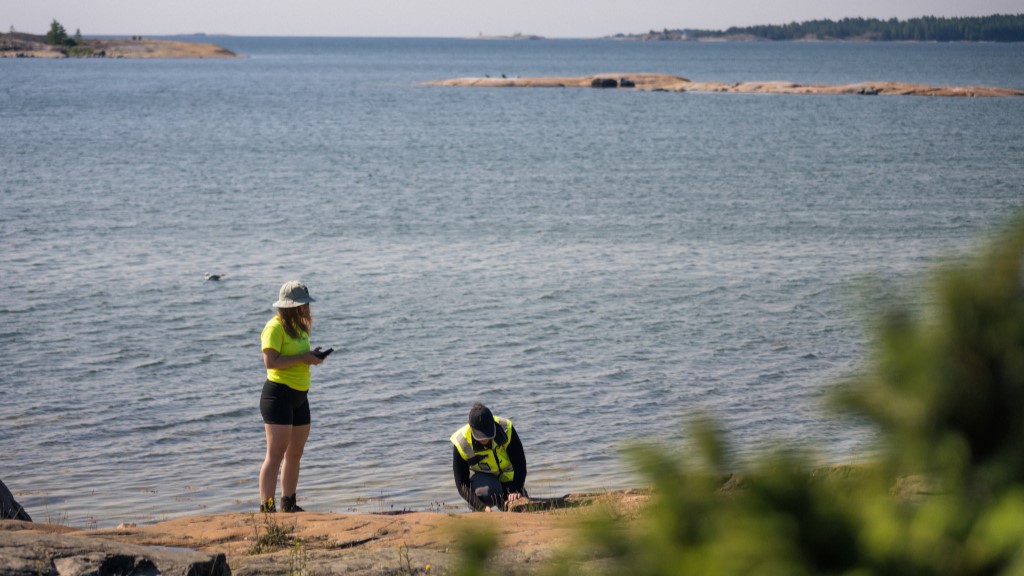 Kaksi henkilöä keltaisissa huomiovaatteissa kalliolla ottamassa näytteitä. Taustalla meri.