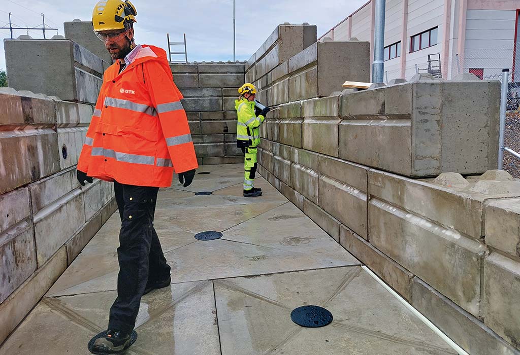 Two workers in high-visibility safety gear inside a concrete-walled test facility. One worker in an orange GTK jacket walks in the foreground, while another in a yellow suit examines the structure in the background.