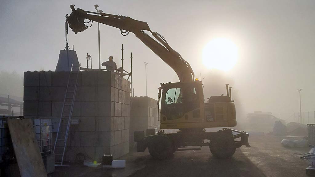 An excavator lifting a large bag onto a concrete block structure at a testing site in foggy conditions, with workers present and the sun shining through the mist